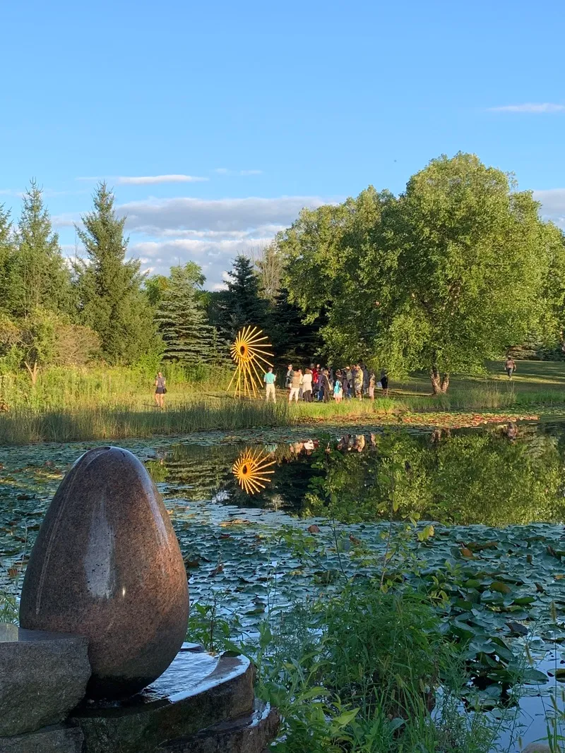 Group of people standing near a yellow sun-shaped sculpture by a pond with lily pads, surrounded by trees under a blue sky.