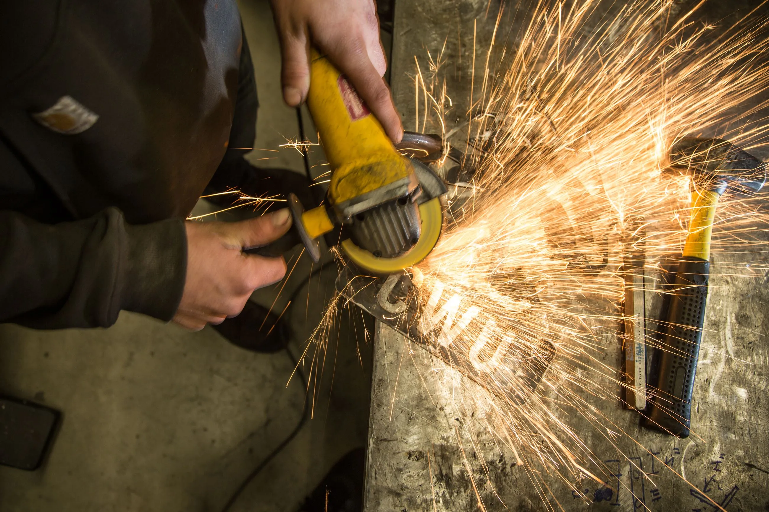 Person using a yellow angle grinder on metal, creating bright sparks on a workbench with a hammer nearby.