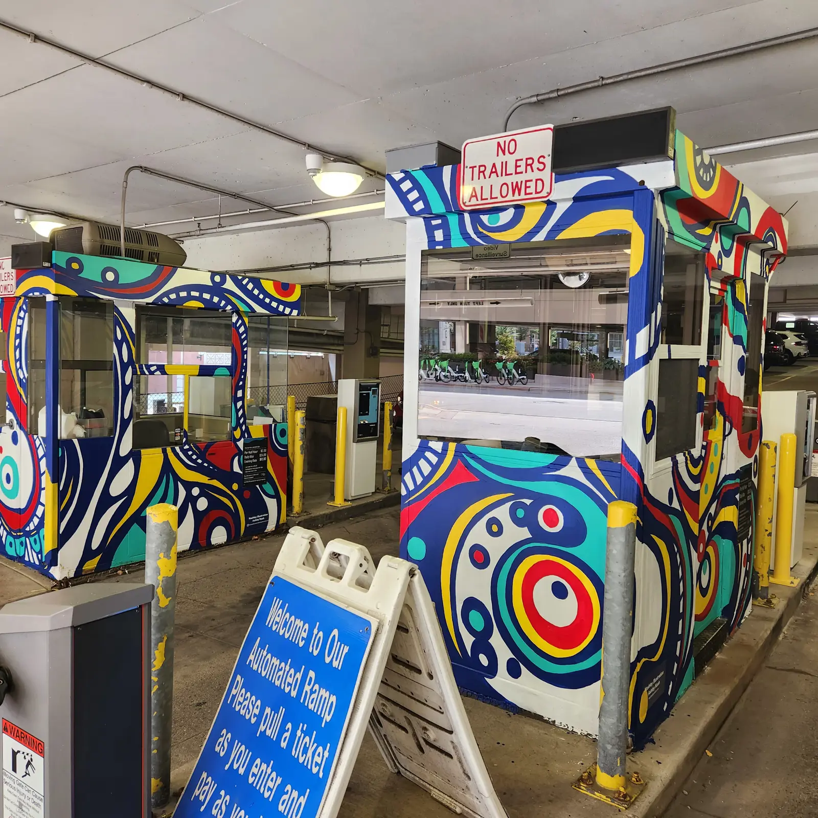 Colorfully painted parking garage pay booths with a sign reading 'No Trailers Allowed' and a blue sign instructing to pull a ticket at the automated ramp entrance.
