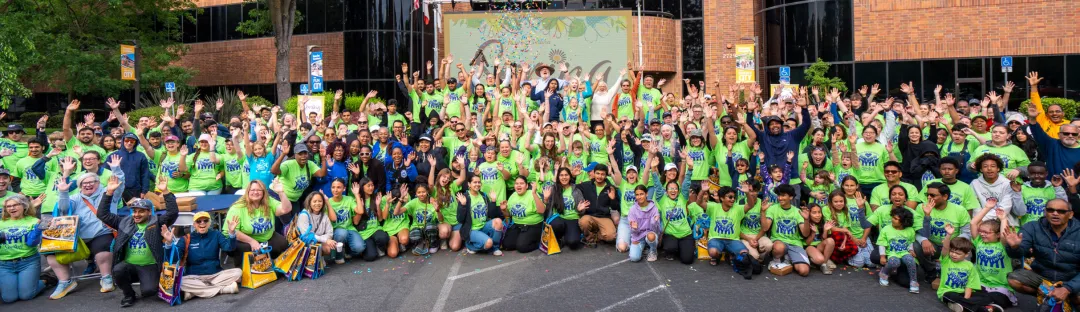 Large diverse group of people wearing bright green event t-shirts gathered outdoors, smiling and waving at the camera.