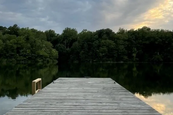 Wooden dock extending over calm lake water with a forested shoreline under a cloudy sky at sunset.