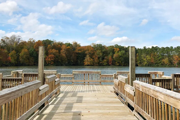 Wooden boardwalk extending over a lake with trees showing autumn colors in the background under a partly cloudy sky.