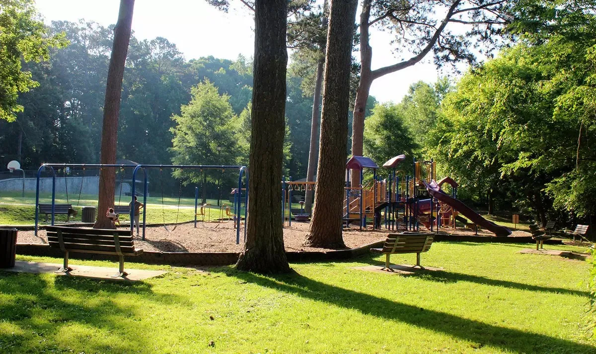 Sunlit playground with swings, slides, benches, and surrounding trees in a green park.