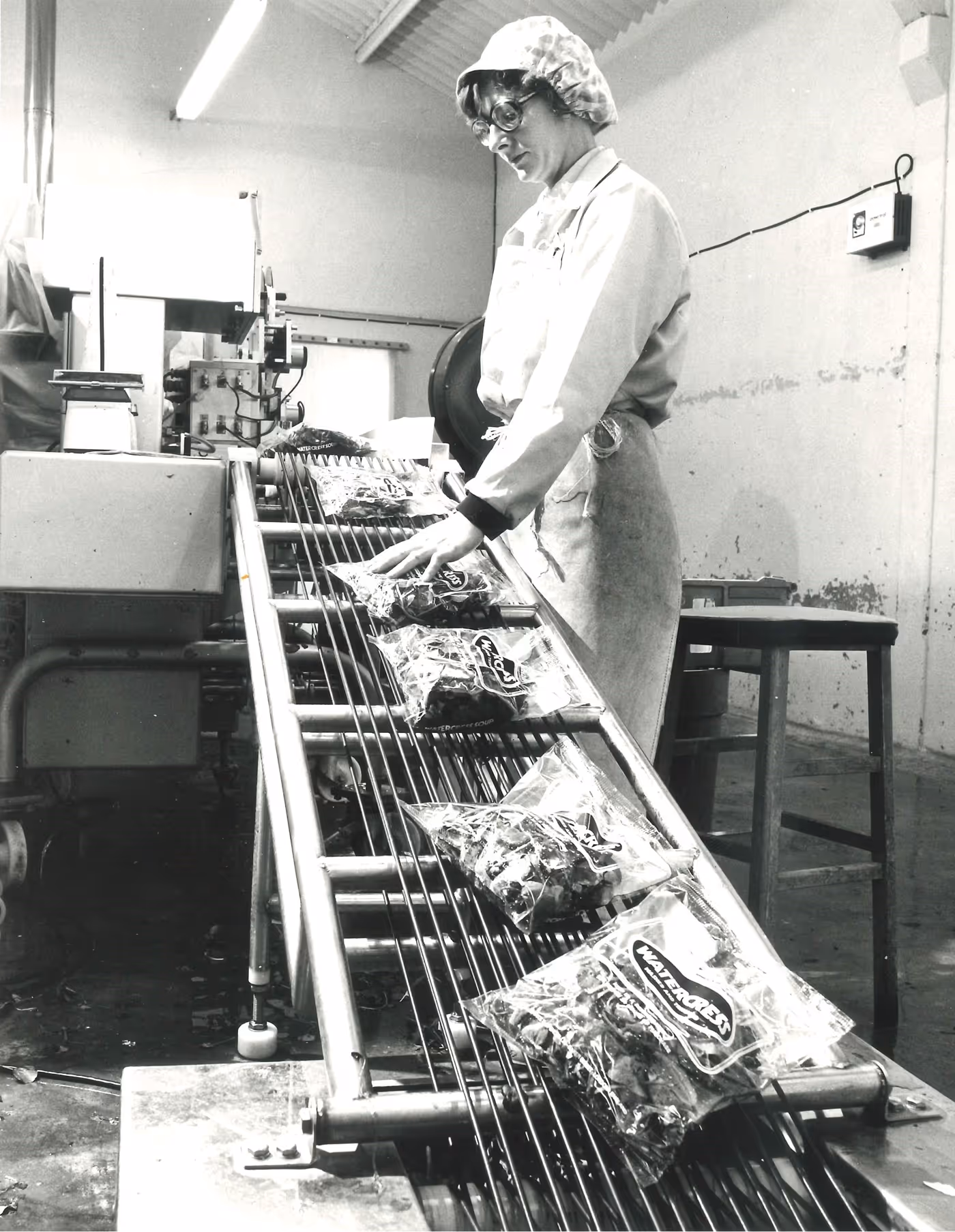 Black and white photo of a worker with glasses and a hairnet inspecting packaged products on a conveyor belt in a factory.