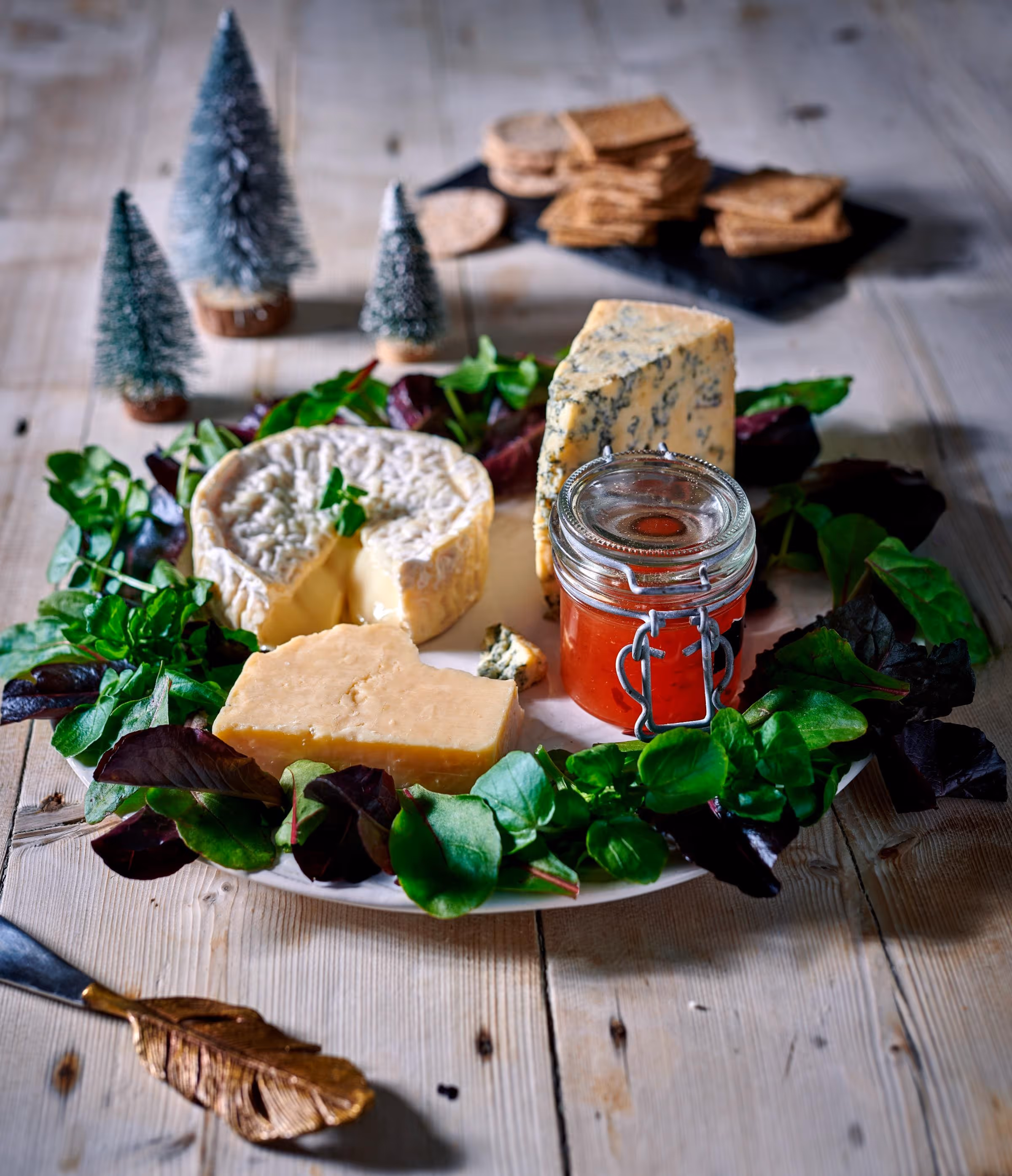 Plate with three types of cheese, a jar of red jam, and mixed greens arranged around the cheese, with crackers and miniature decorative trees in the background.