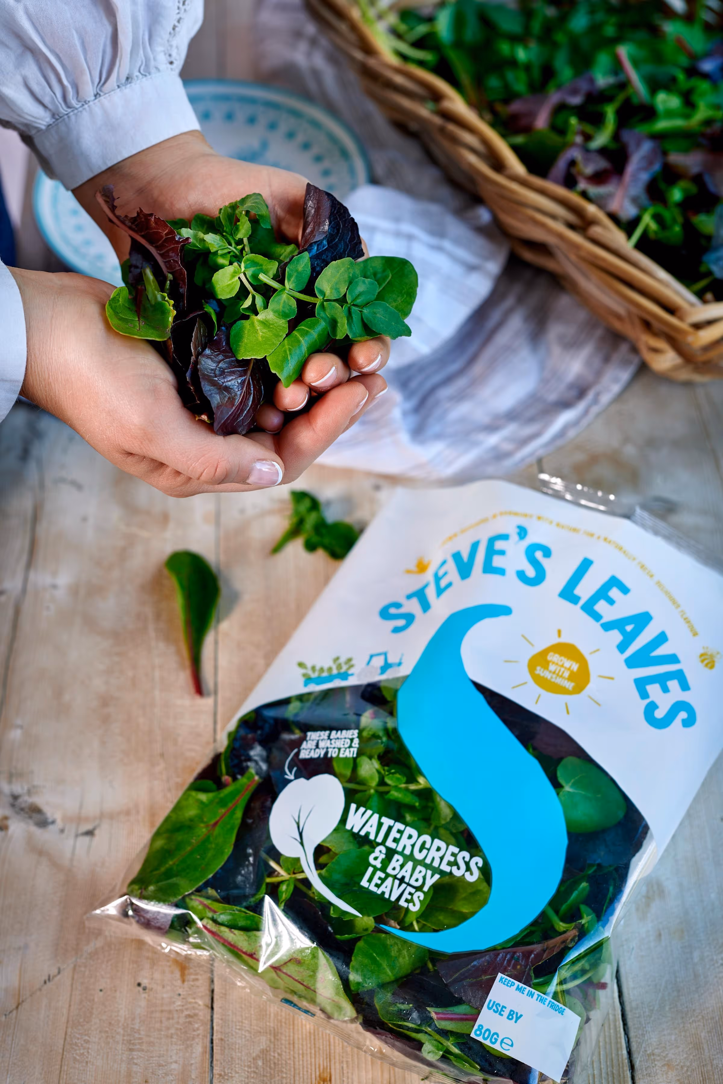 A person holding fresh watercress and baby leaves above a wooden table with a bag of Steve's Leaves mixed salad.