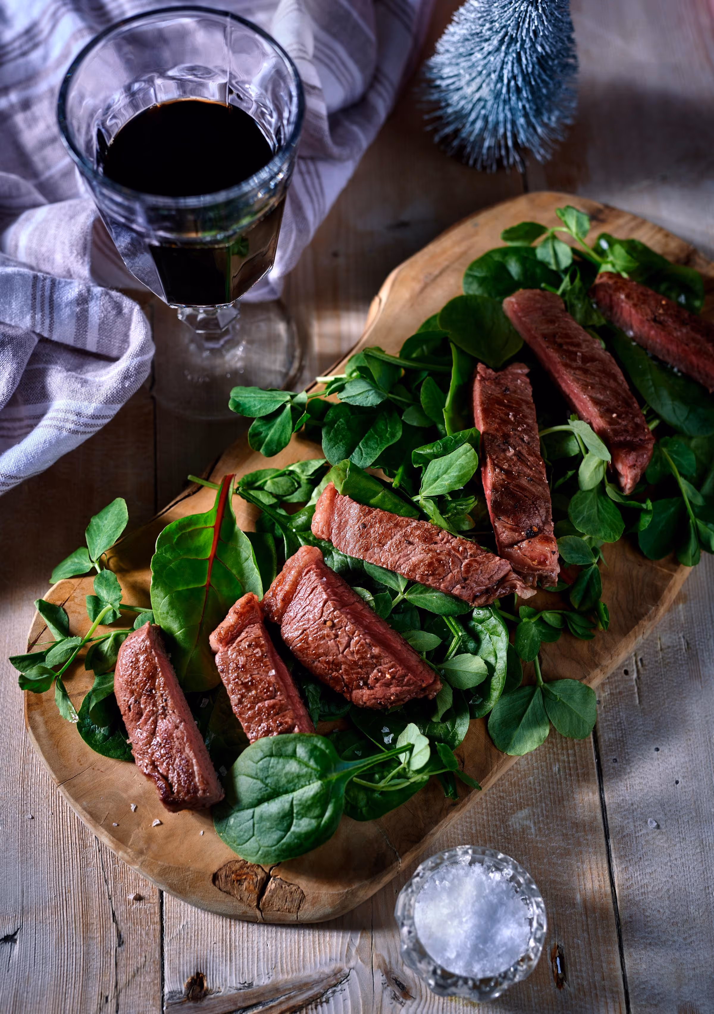 Slices of cooked steak served on fresh spinach and greens on a wooden cutting board next to a glass of red wine and a salt container.
