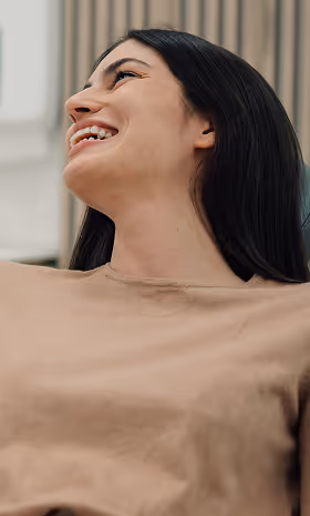 Smiling woman with long dark hair looking up, wearing a beige top indoors.