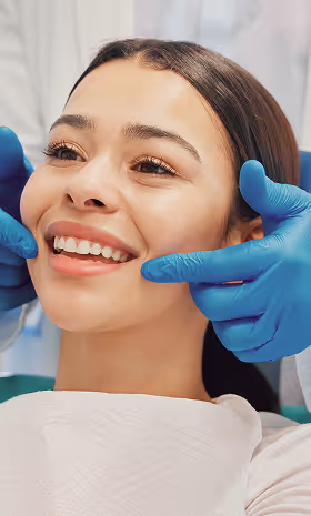 Dentist wearing blue gloves examining a young woman smiling in dental chair.