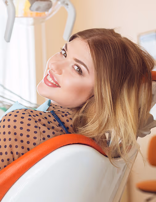 Smiling woman with light brown hair reclines in an orange and white dental chair at a clinic.