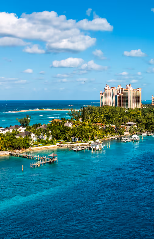 Coastal view with turquoise water, docks, greenery, houses, and tall buildings under a blue sky with clouds.