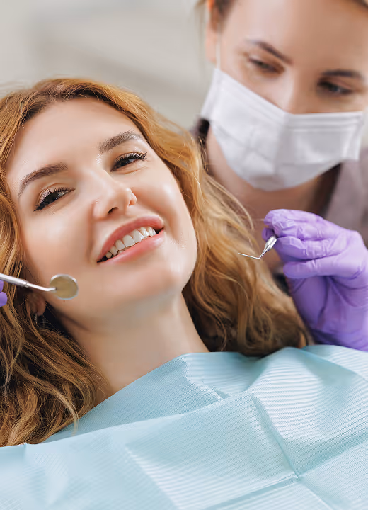 Female dentist wearing mask and gloves examining smiling woman's teeth with dental tools.