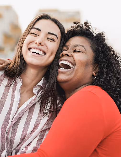 Two women smiling and laughing together outdoors, one wearing a striped shirt, the other a red top.