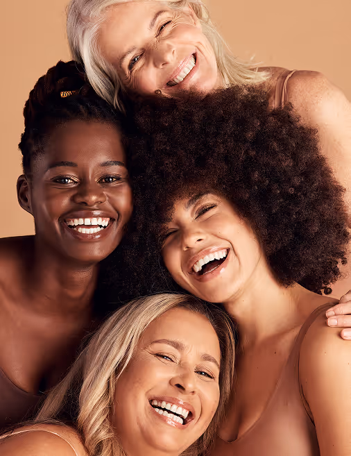 Four diverse women smiling and embracing each other against a beige background.