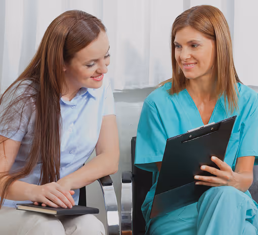 Two women seated indoors, one in medical scrubs holding a clipboard, both engaged in conversation.