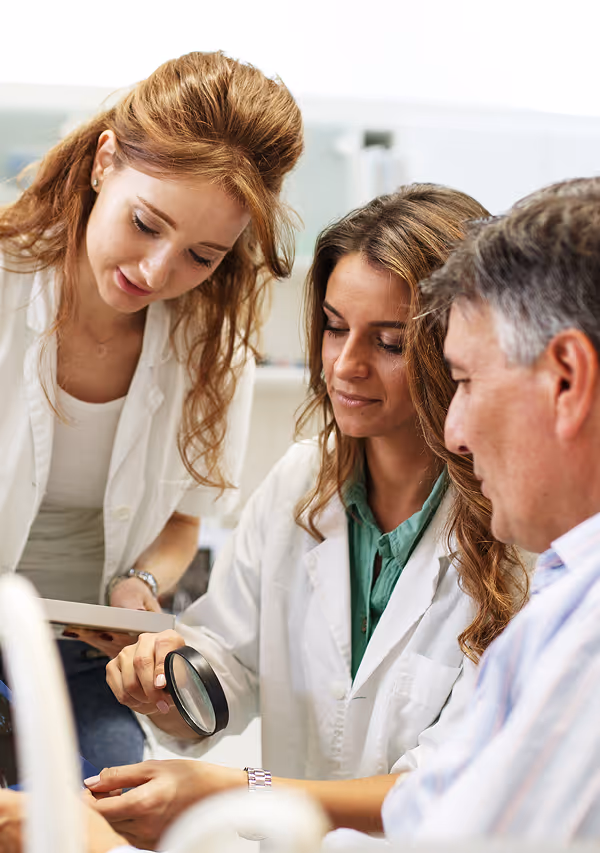 Two women in lab coats examine a patient's skin using a magnifying glass while a man watches closely.