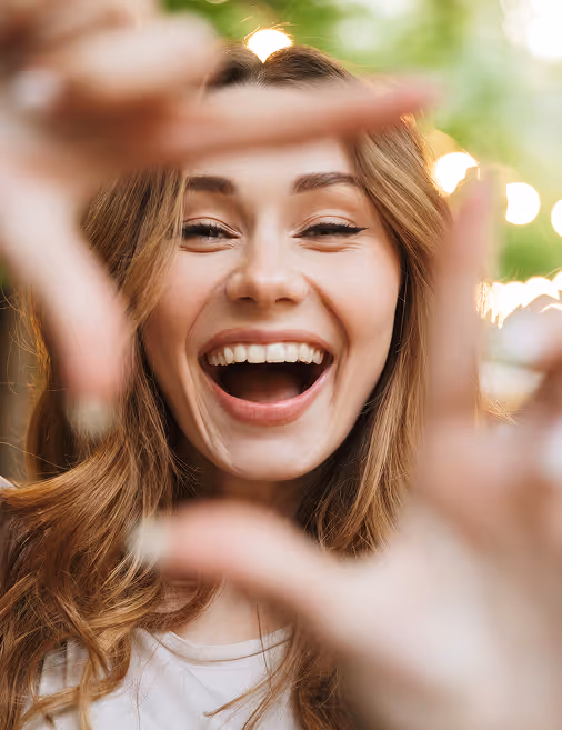 Happy young woman smiling and framing face with her hands outdoors with blurred lights behind.
