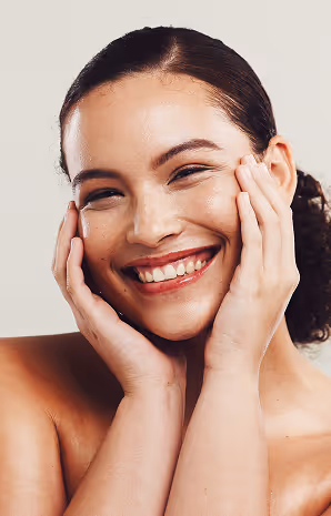 Smiling woman with clear skin holding her face with both hands against a plain background.