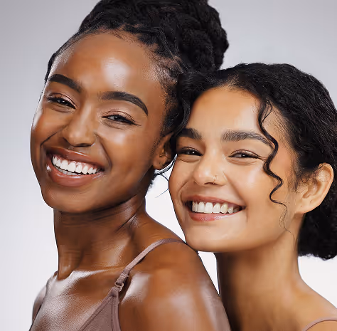 Two smiling women with smooth skin, one with dark braids and the other with curly hair and a nose ring.