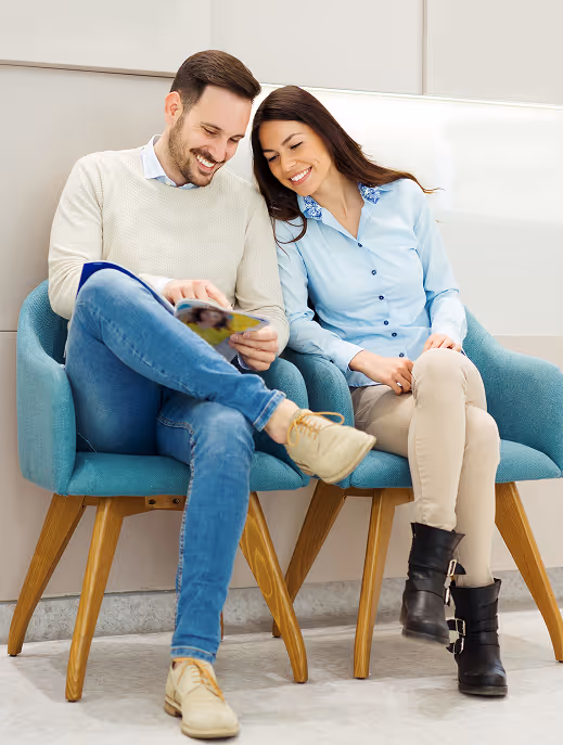 Smiling man and woman sitting on blue chairs, reading a magazine together in a light room.