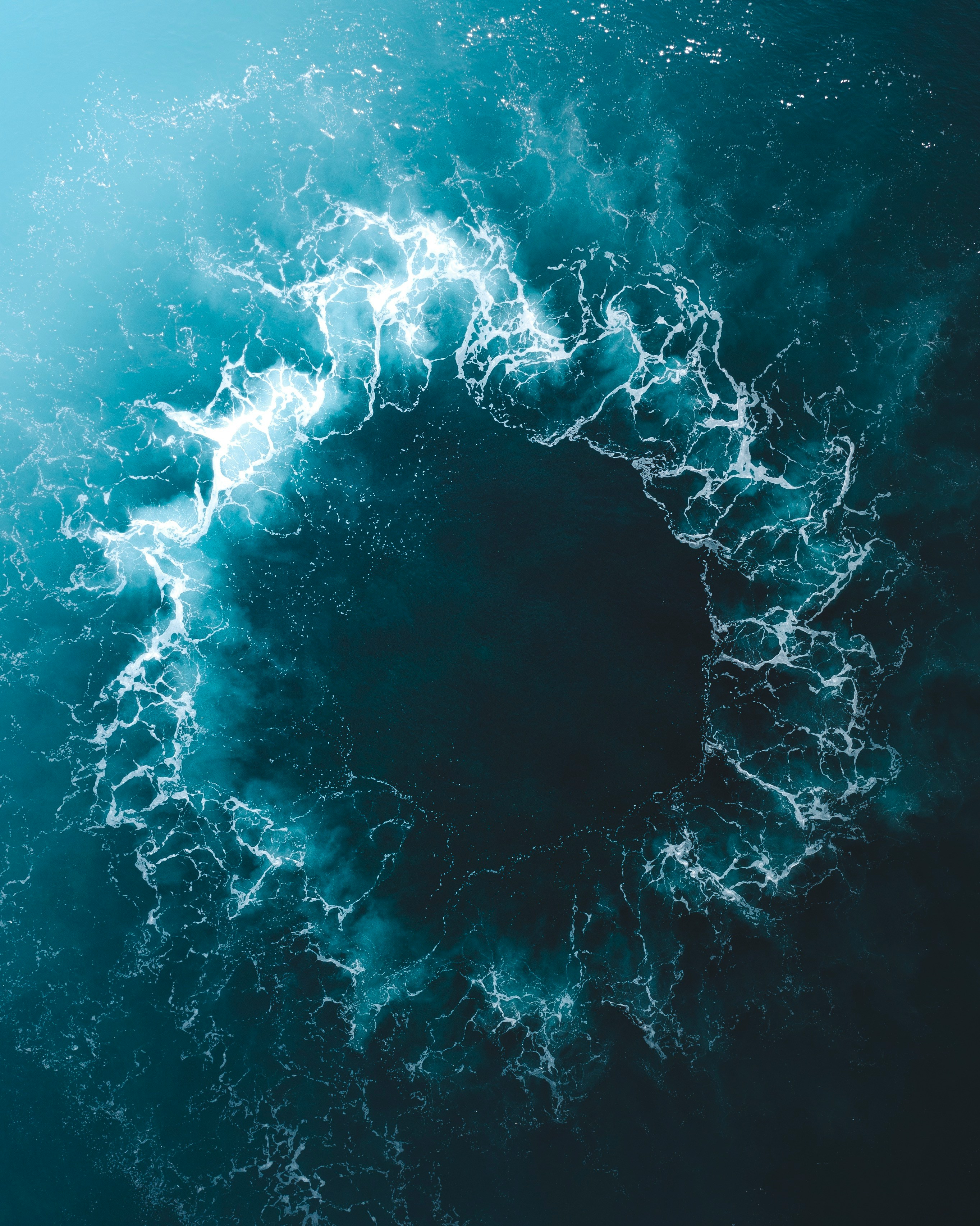 Aerial view of ocean water swirling in a circular pattern with white foam on deep blue surface.