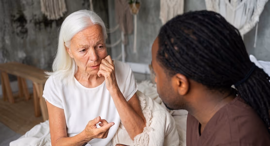 Elderly white woman looks concerned while talking to a man with braided hair indoors.