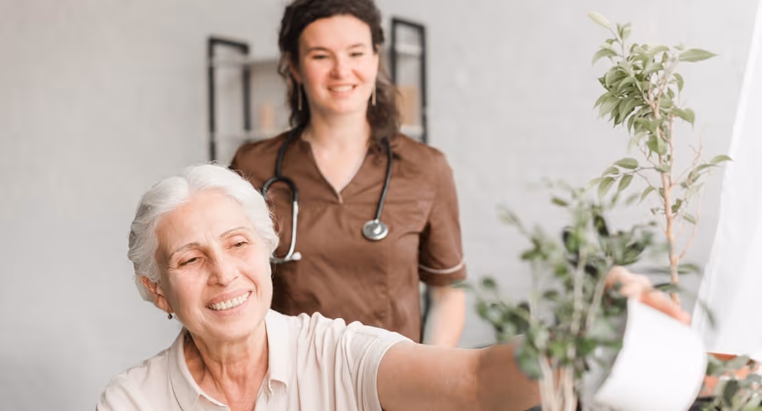 Smiling elderly woman watering a plant with a young female nurse standing behind her.