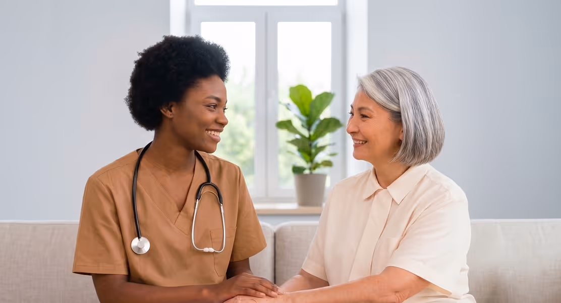 Smiling female doctor in scrubs holding hands with elderly woman sitting on a couch together.