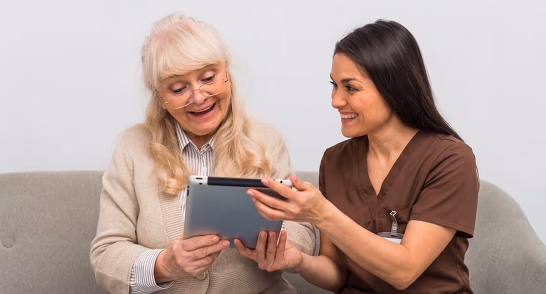 Smiling healthcare worker shows tablet to happy elderly woman sitting on couch together.