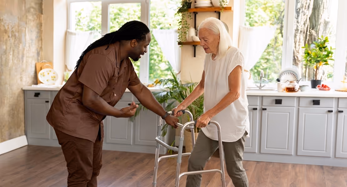 Caregiver assisting elderly woman using a walker in a bright kitchen with plants and large windows.