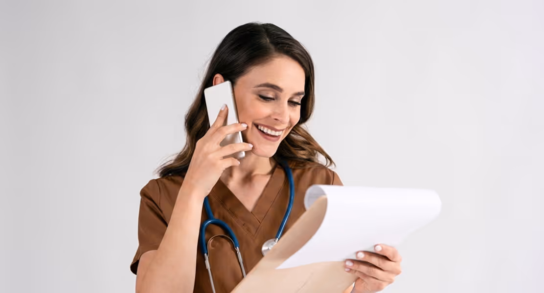 Smiling female healthcare worker with stethoscope talking on phone and reviewing papers.