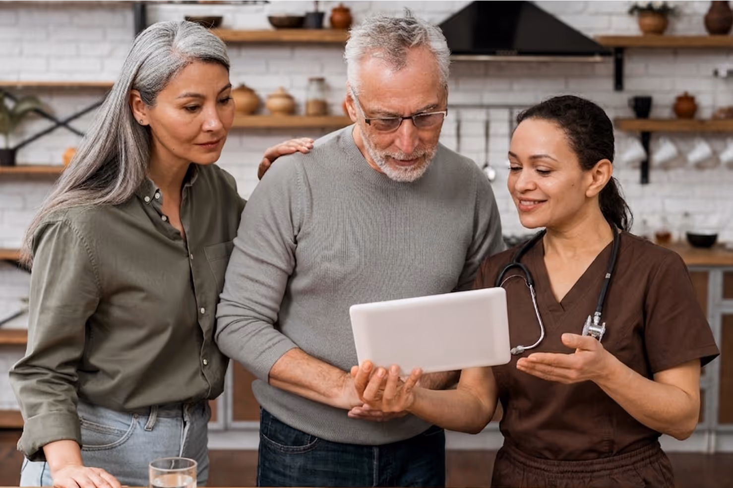 Medical professional shows tablet to an elderly couple in a kitchen setting.