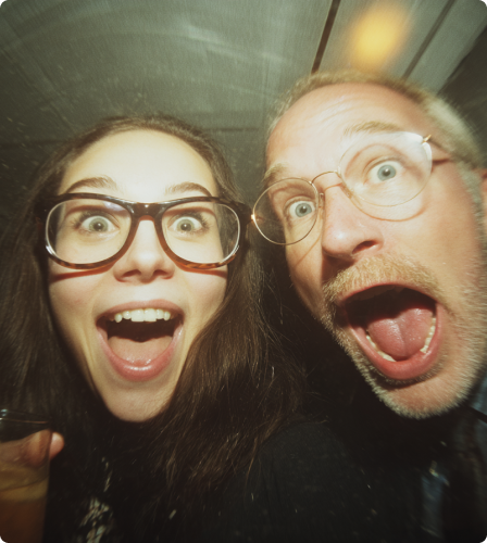 Close-up of a joyful woman and an older man with glasses making surprised, excited faces at the camera.