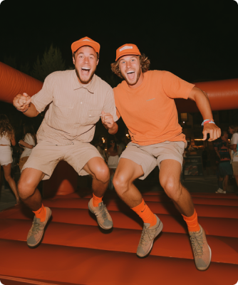 Two young men in shorts and caps jumping excitedly on a red inflatable bounce house at night.