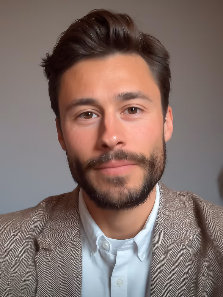 Portrait of a man with short brown hair and beard wearing a brown herringbone blazer and white shirt against a plain background.
