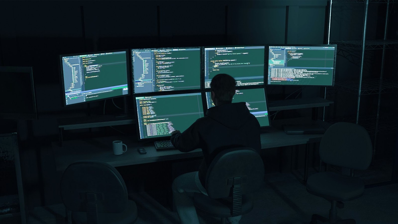 Person sitting at a desk working on multiple computer screens displaying programming code in a dark room.