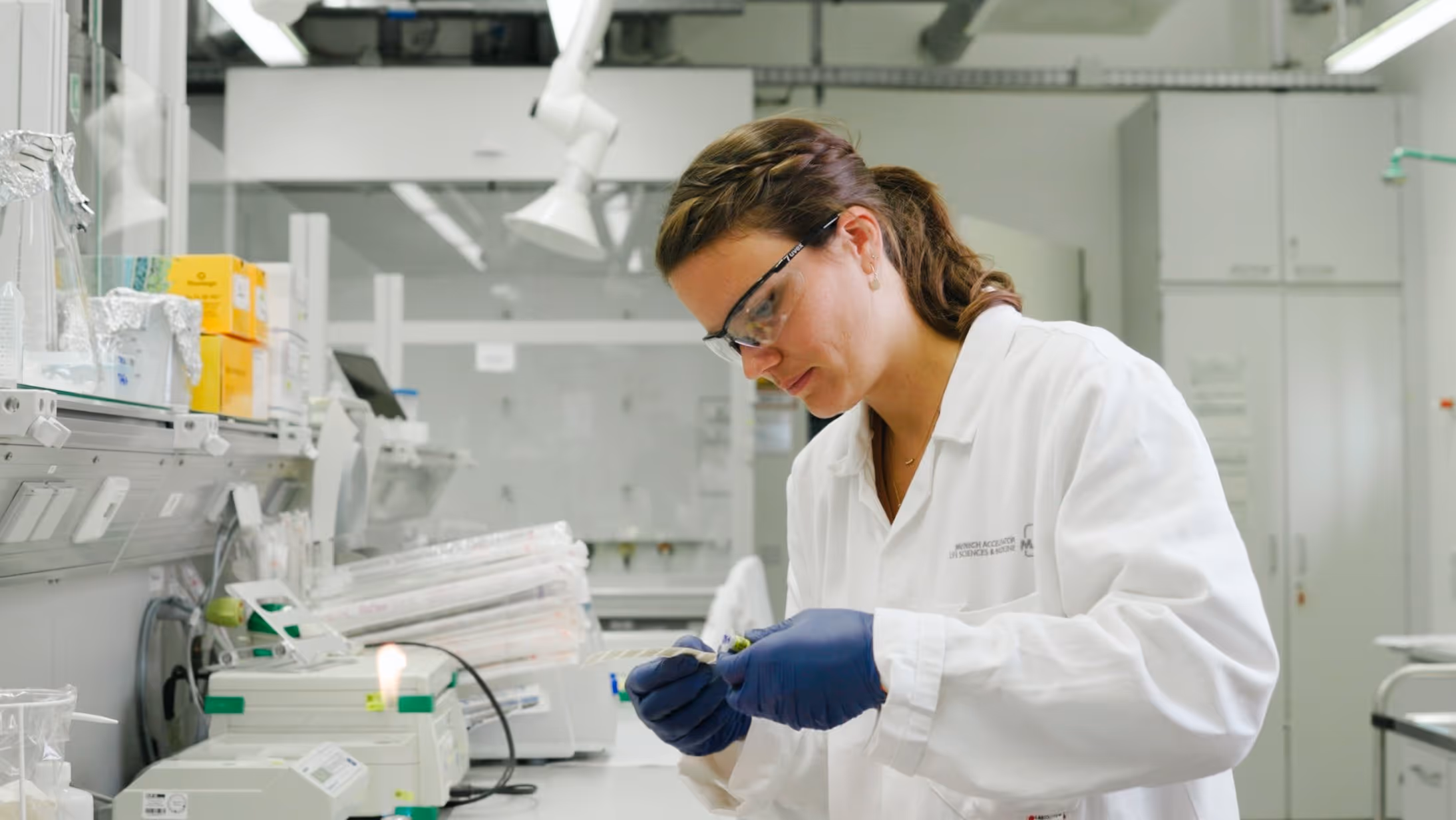 Scientist wearing a white lab coat, safety glasses, and blue gloves working with a sample in a laboratory.