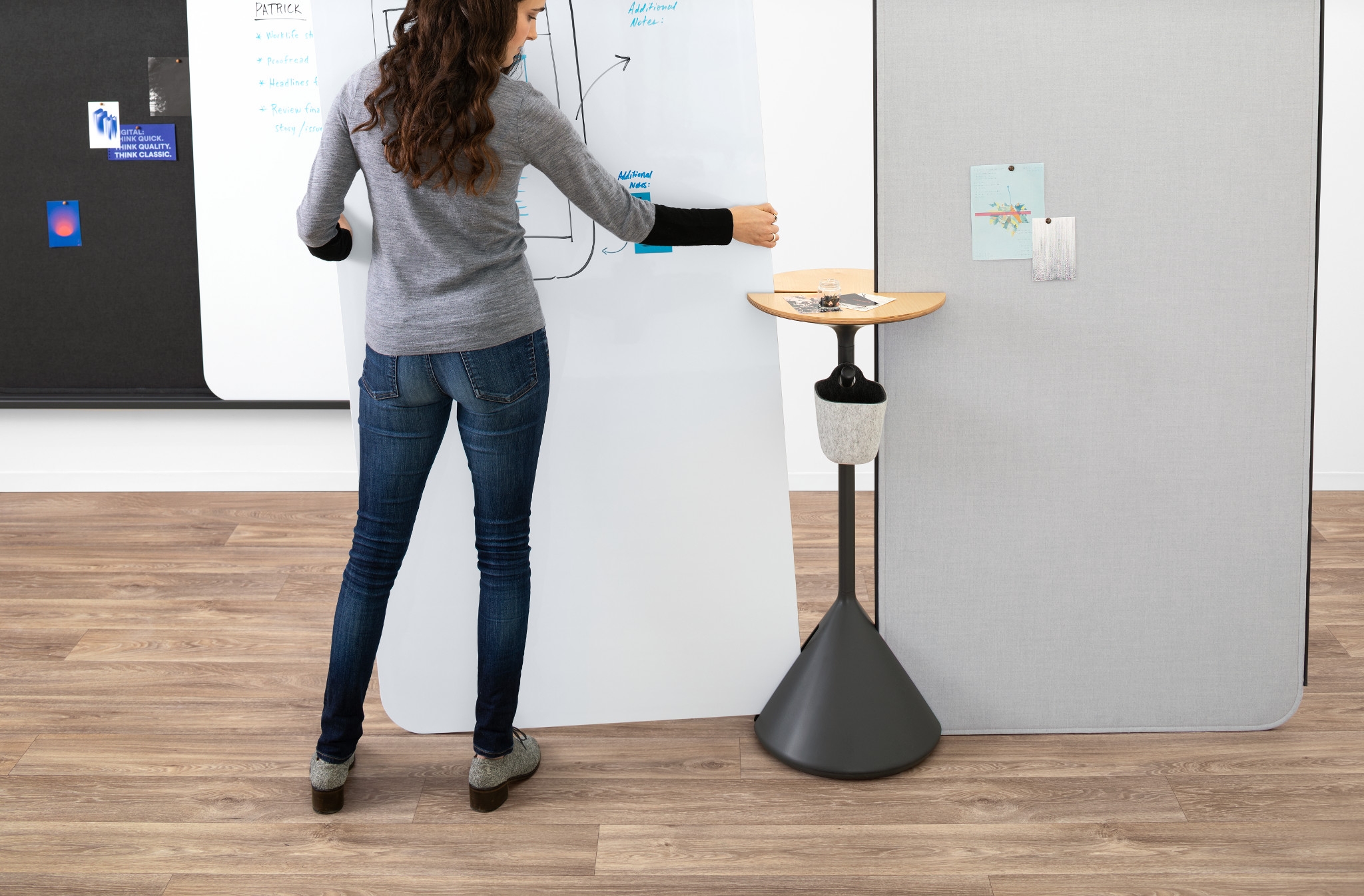 Woman in jeans and gray sweater writing on a large whiteboard with notes and diagrams in a modern office space.