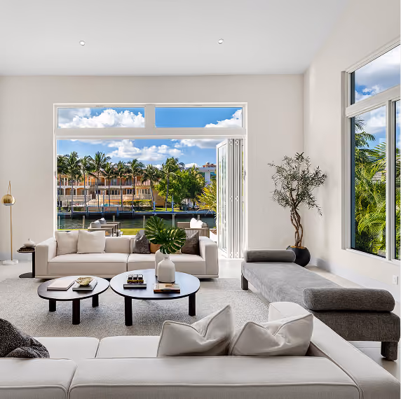 Modern living room with beige sofas, round black tables, and open view of palm trees and waterfront houses.