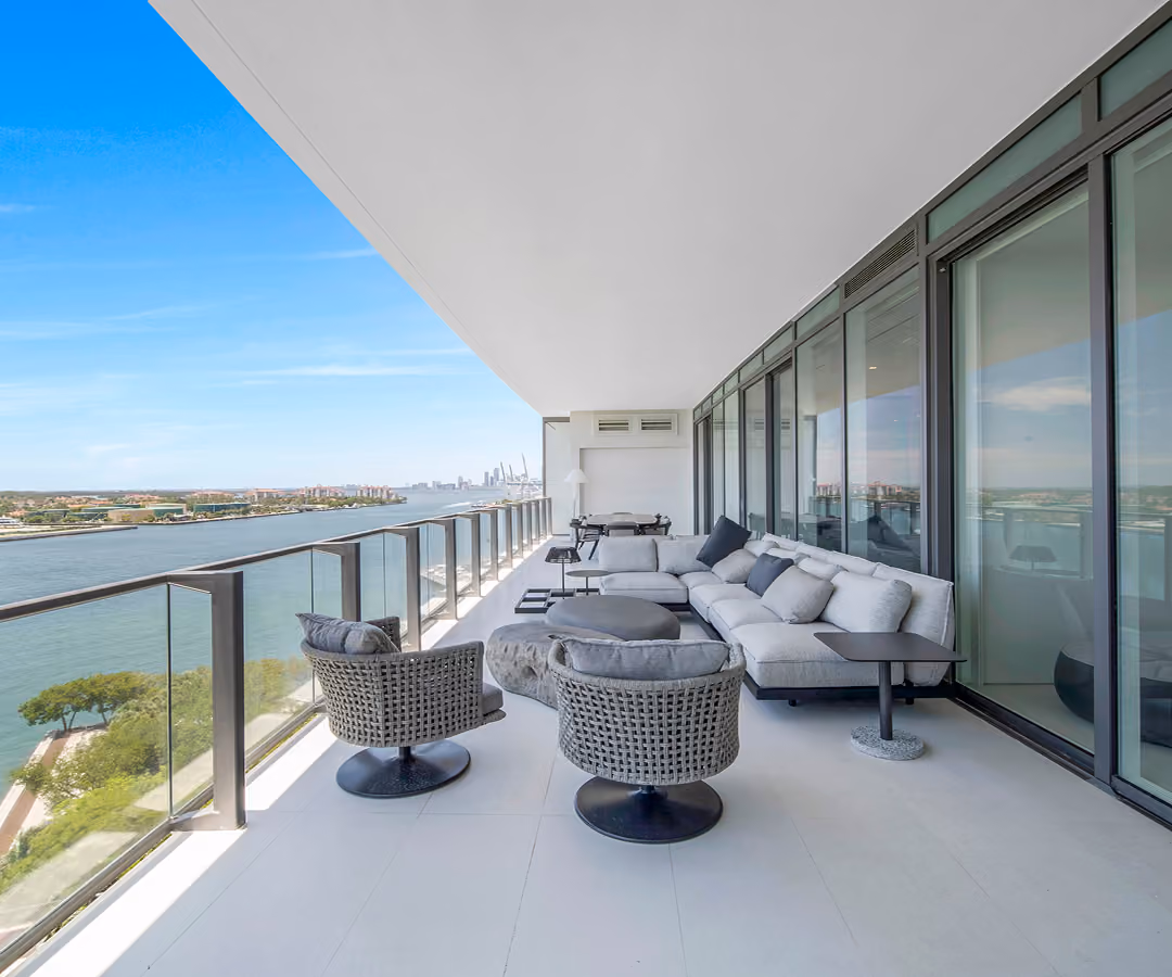 Spacious balcony with woven chairs, cushioned sofa, side tables, and water view under blue sky.