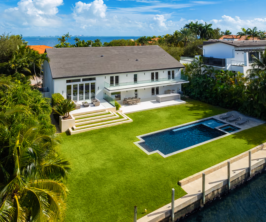 Modern waterfront house with large lawn, rectangular pool, palm trees, and ocean view under blue sky.
