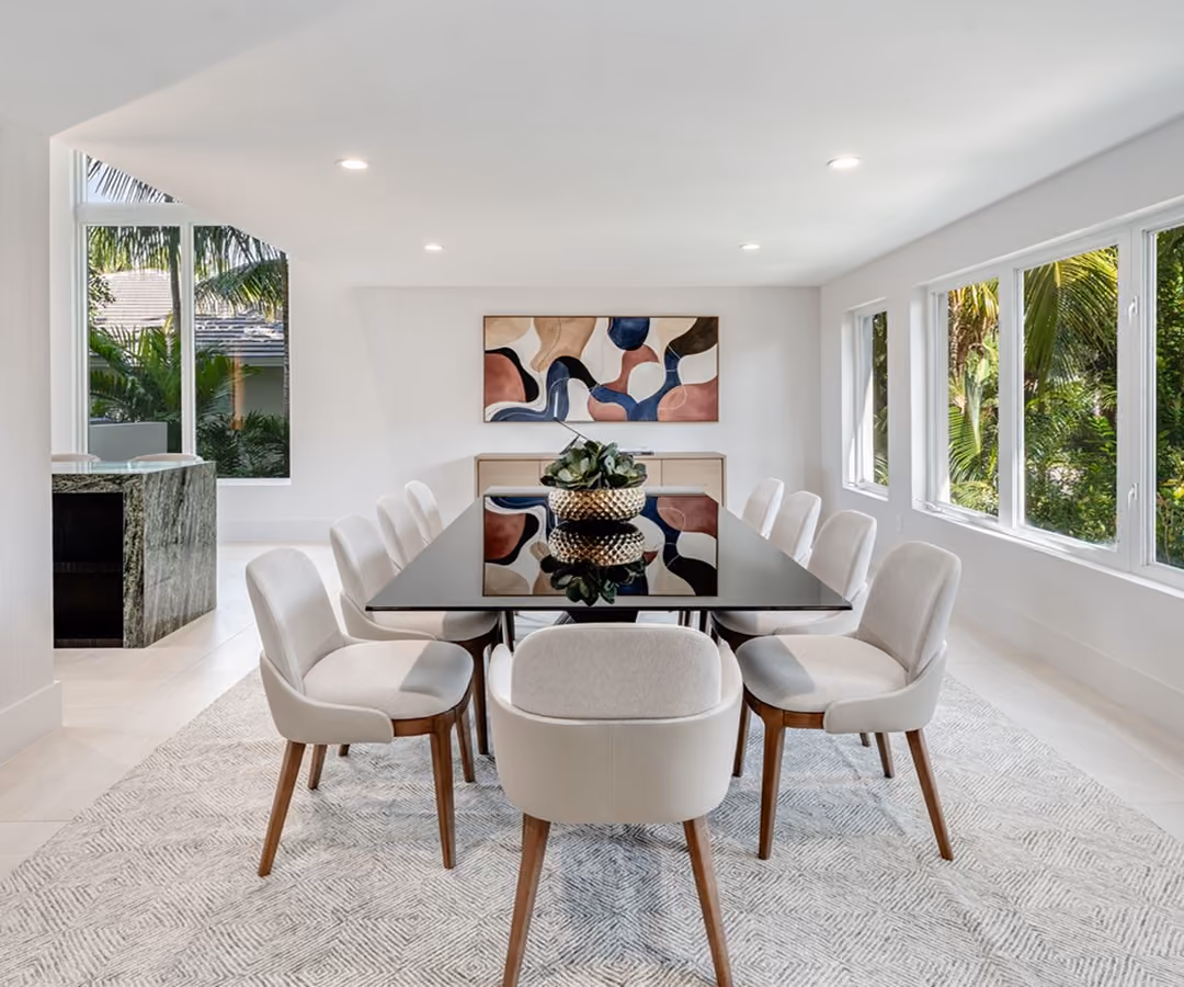 Modern dining room with black table, ten beige chairs, abstract wall art, and large windows with greenery outside.