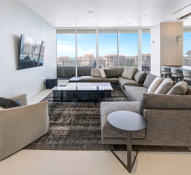 Modern living room with large gray sofas, a wall-mounted TV, glass coffee table, and floor-to-ceiling windows.