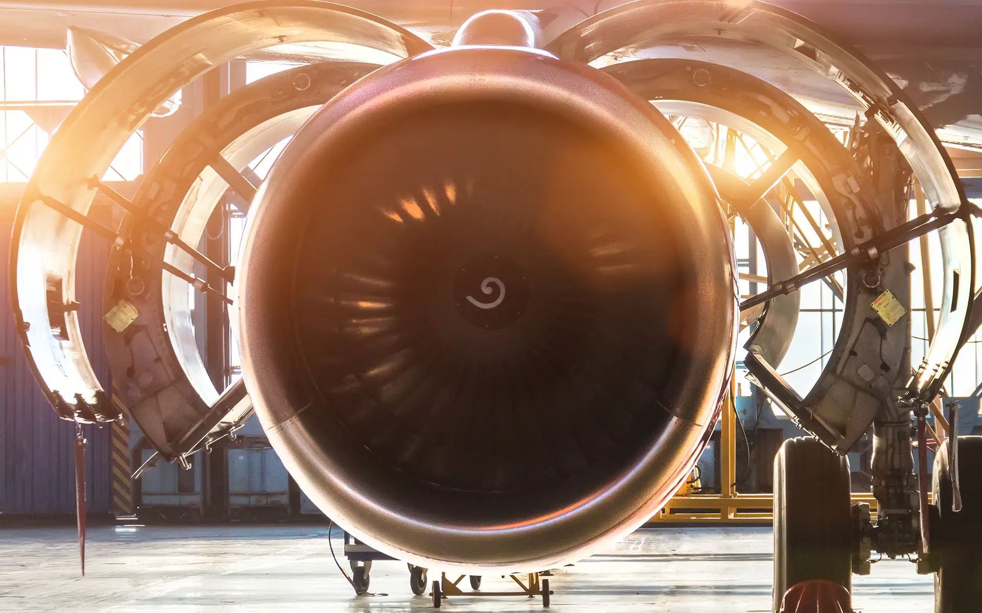 Close-up front view of a large aircraft jet engine with its cowling open inside a hangar illuminated by sunlight.