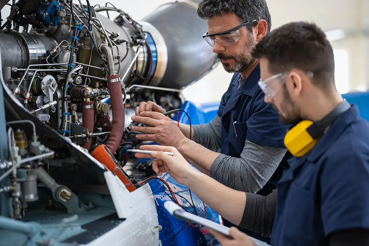 Two engineers wearing safety glasses inspecting and working on an aircraft engine with diagnostic equipment.