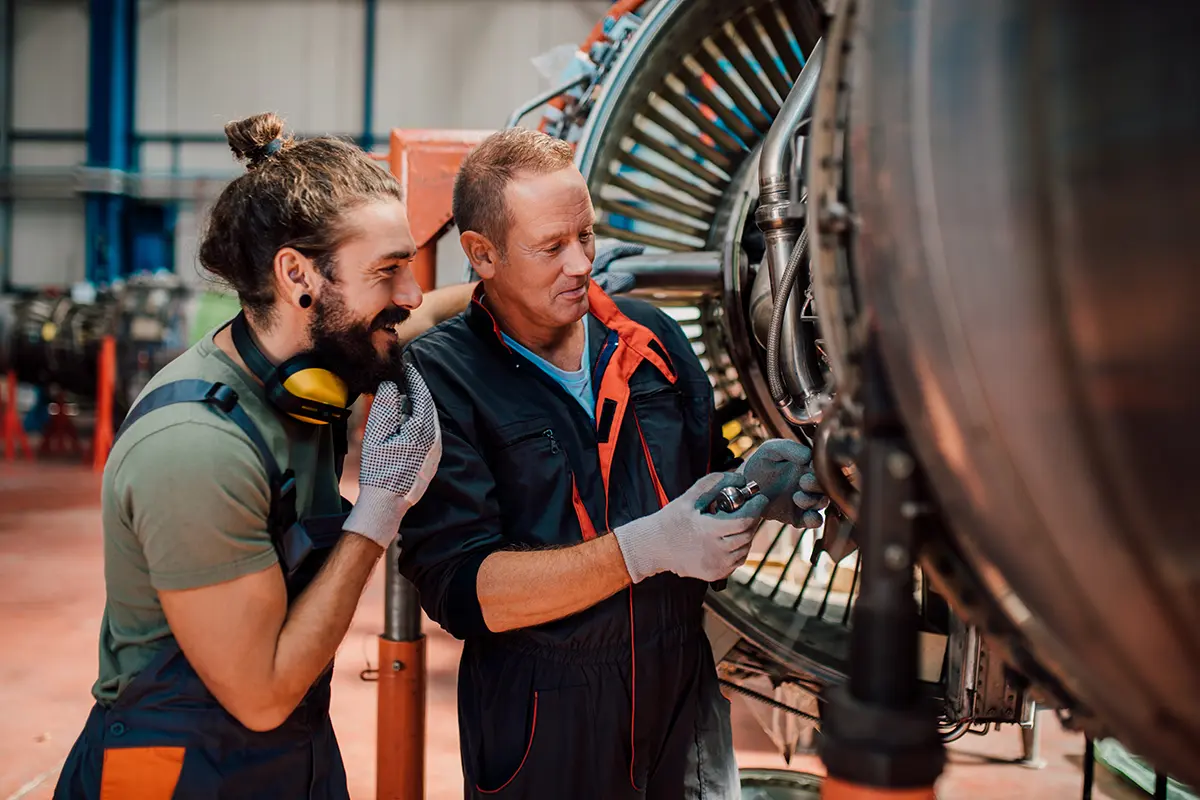 Two mechanics training and working together inspecting and repairing a turbine engine in an industrial workshop.