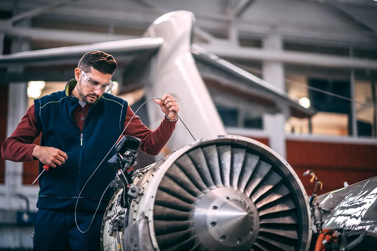 Technician wearing protective glasses inspecting and testing an aircraft jet engine inside a hangar.
