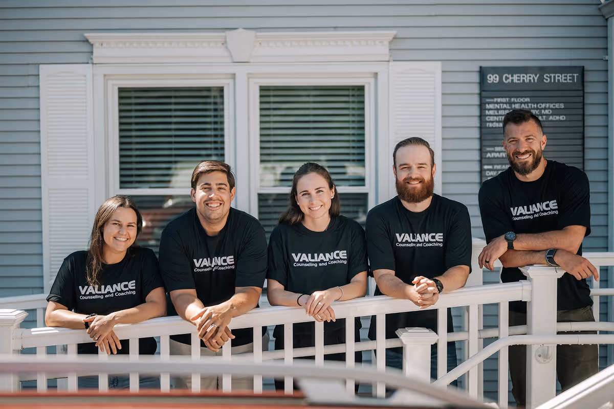 Five people in black Valiance Counseling and Coaching shirts smiling on porch railing outside a blue building.
