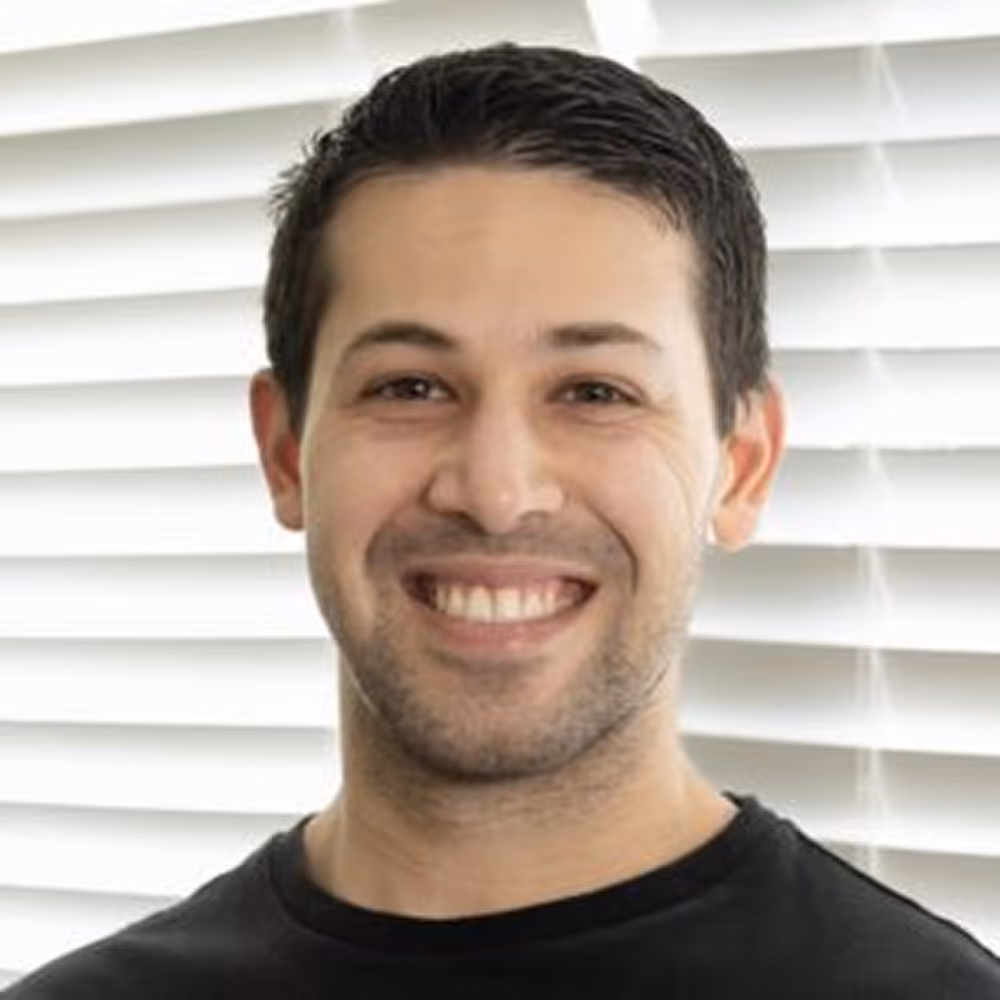Smiling man with short dark hair wearing a black shirt, standing in front of white blinds.