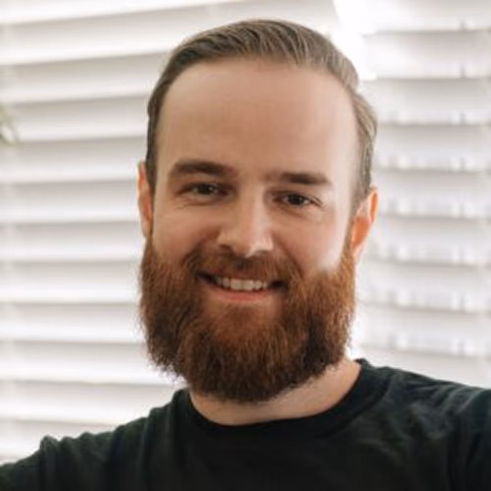 Smiling man with a full reddish beard and slicked-back hair wearing a black shirt in front of white blinds.